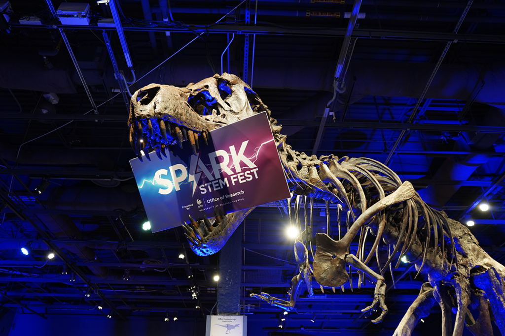 A dinosaur skeleton holds a "Spark STEM Fest" sign in its mouth inside an exhibition space with blue lighting.