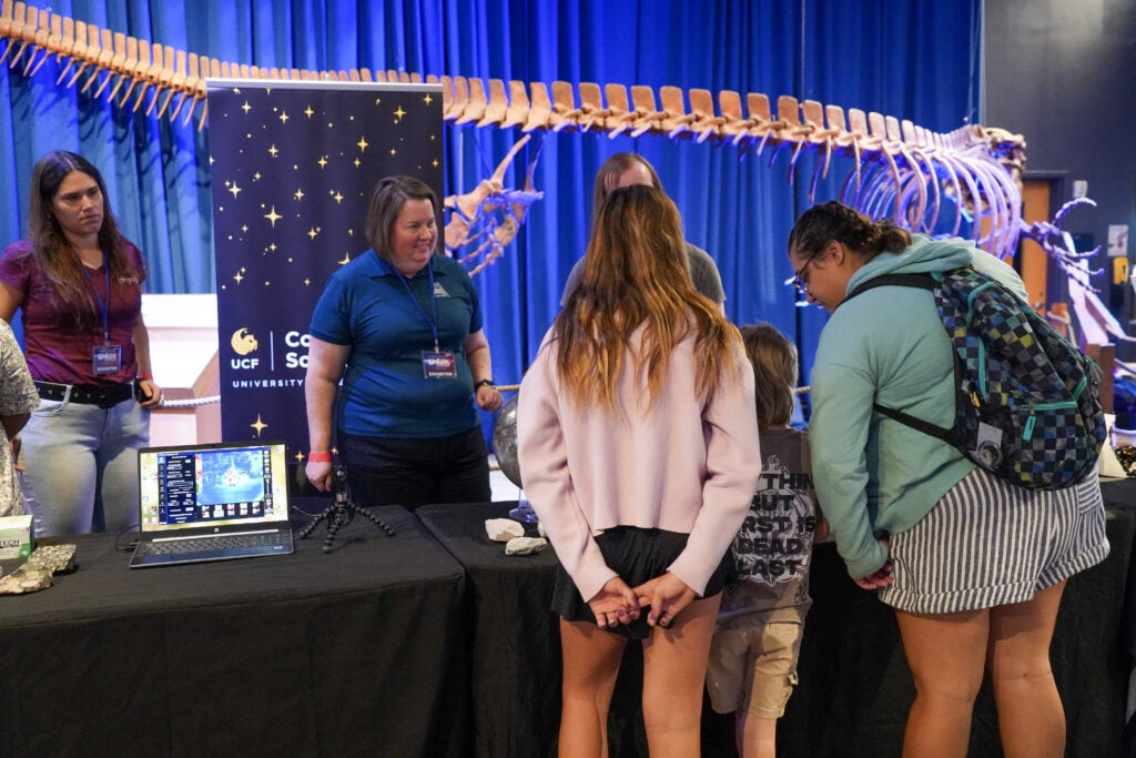 A group of people interact at an educational booth in a museum, with a large dinosaur skeleton displayed in the background.