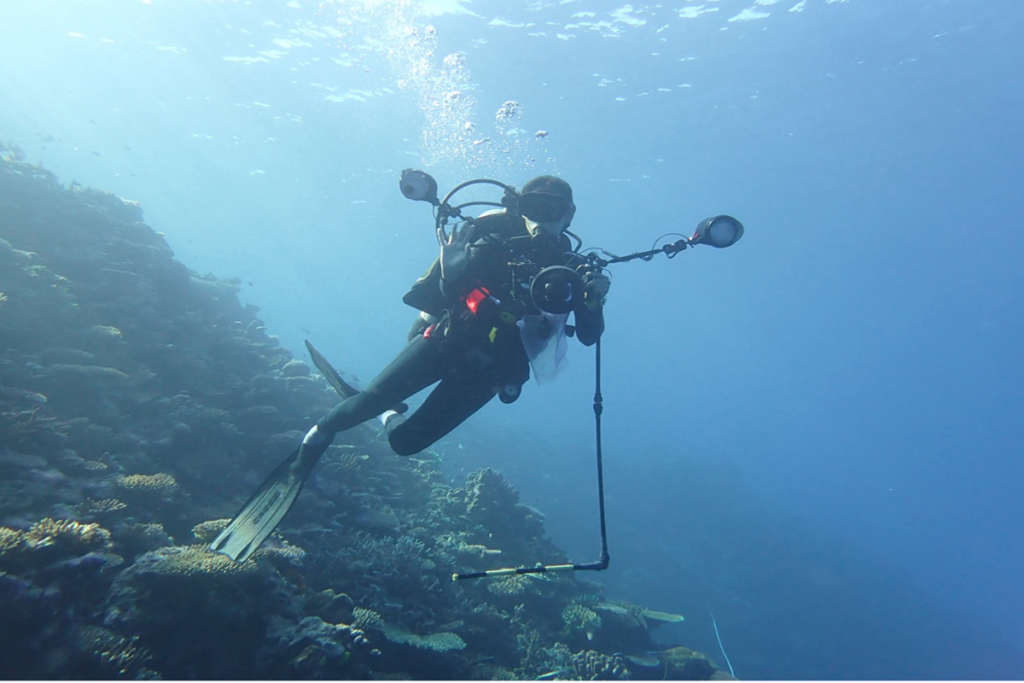 A scuba diver equipped with underwater photography gear swims above a coral reef in clear blue water.