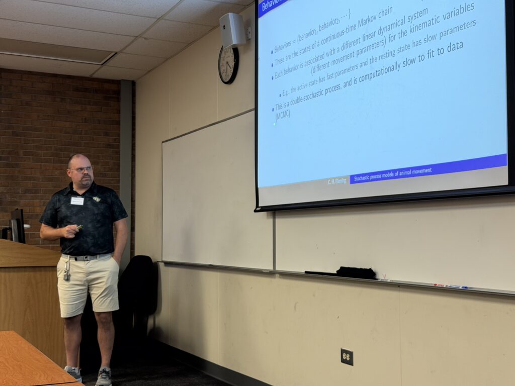A man stands near a projector screen in a classroom, presenting a slide on Markov chains and stochastic processes to an audience.