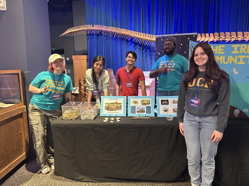 Five people stand behind and beside a display table with educational materials and specimens at an indoor event; a blue curtain and large skeleton are visible in the background.