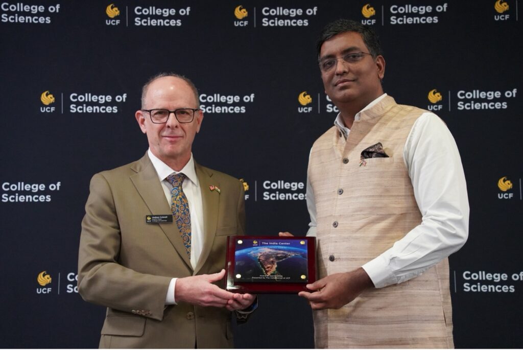 Two men stand in front of a UCF College of Sciences backdrop; one hands the other a plaque featuring an image of India.