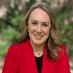 Woman with wavy blonde hair wearing a red blazer and black top, smiling outdoors with greenery in the blurred background.