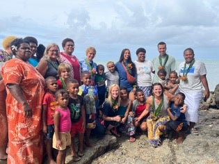 A diverse group of adults and children pose together on rocky ground near the ocean under a cloudy sky.