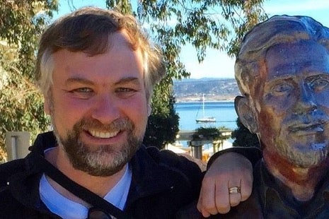 A man stands outdoors smiling next to a bronze statue, with trees, water, and a sailboat visible in the background.