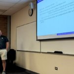 A man stands near a whiteboard giving a presentation in a classroom; a PowerPoint slide about Markov chains is projected on the wall.