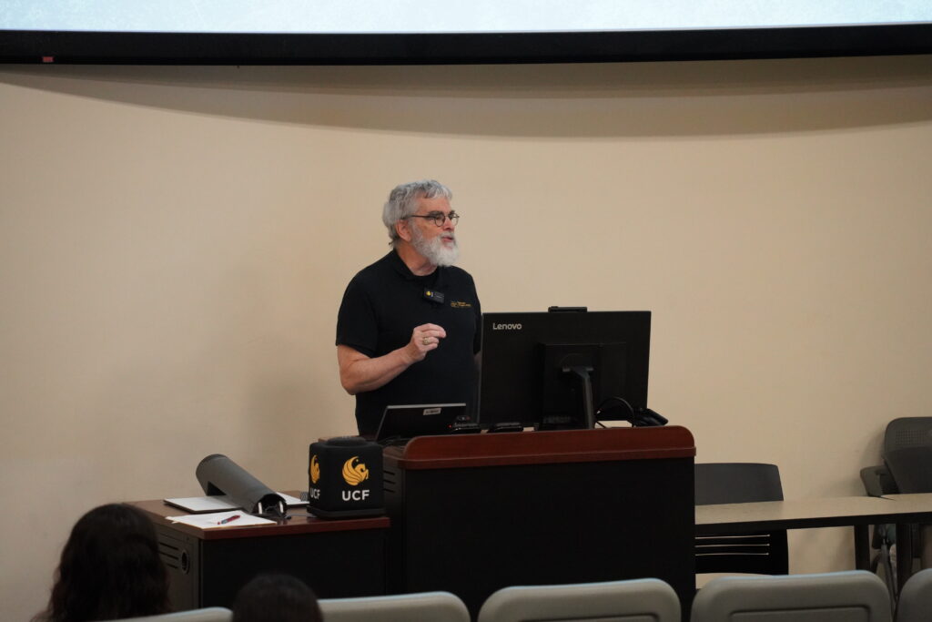A man with gray hair and beard speaks at a podium in a lecture hall, standing behind a computer with a UCF-branded box on the desk.