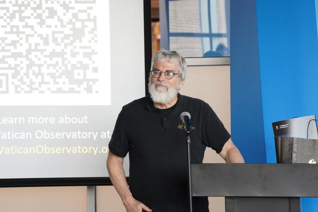 An older man with gray hair and a beard stands at a podium, speaking into a microphone. A presentation screen with a QR code and text is visible in the background.