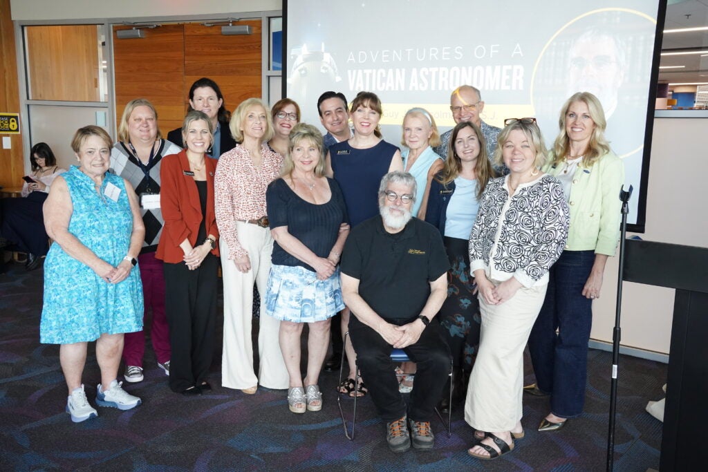 A group of people poses together indoors in front of a projection screen displaying "Adventures of a Vatican Astronomer." One person is seated while the rest stand around him.