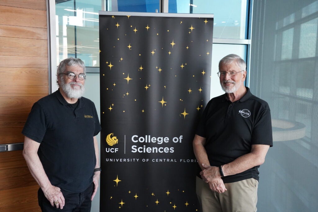 Two older men stand on either side of a UCF College of Sciences banner inside a modern building with glass and wood paneling.