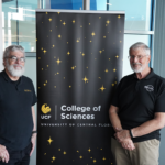 Two men stand beside a University of Central Florida College of Sciences banner, posing for a photo indoors.