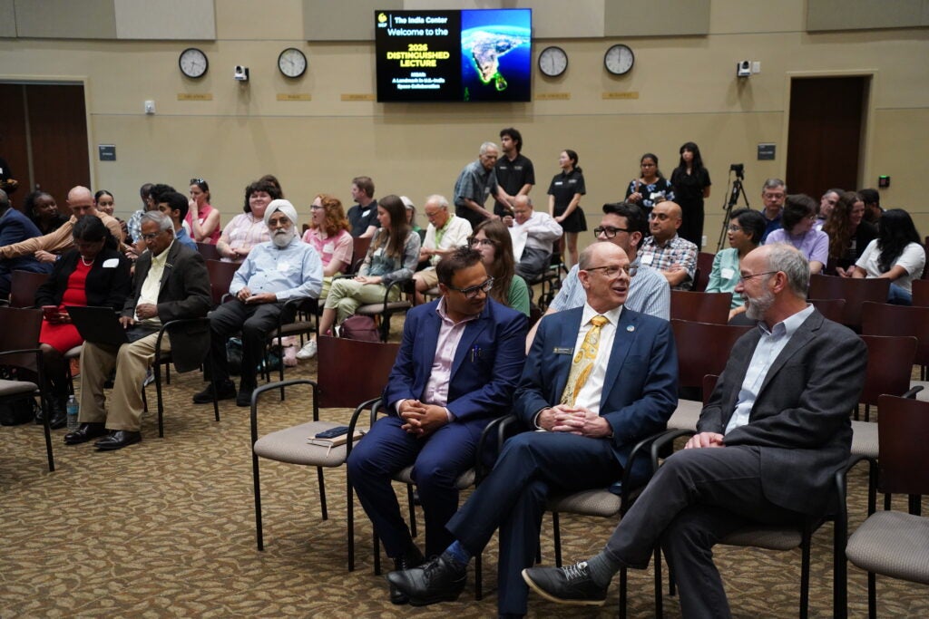 A group of people seated in an auditorium, some talking among themselves, with a presentation screen and clocks visible on the wall behind them.