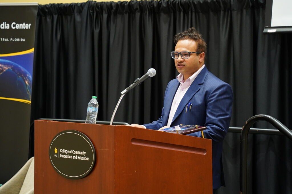 A man in a suit speaks at a podium with a microphone and water bottle. The podium displays the College of Community Innovation and Education logo. A black curtain is in the background.