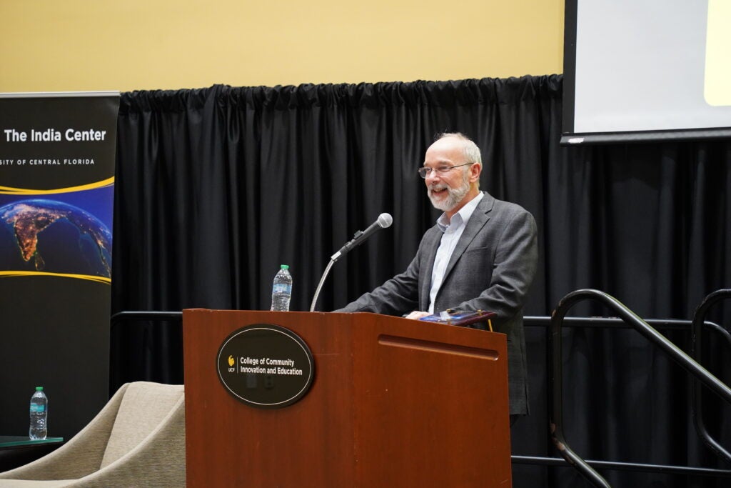 A man in a suit speaks at a podium labeled "College of Community Innovation and Education" with a water bottle and books on the podium. A banner for The India Center is visible in the background.