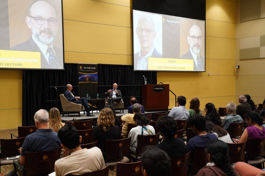 Two speakers sit on stage in armchairs for a lecture, addressing an audience; large screens behind them display a portrait and event details about space collaboration.