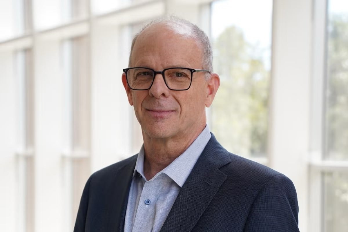 A man wearing glasses and a dark suit jacket stands indoors in front of large windows with natural light.