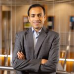 A man in a gray suit stands indoors with arms crossed, smiling at the camera. The background features blurred lights, railings, and wall-mounted frames.