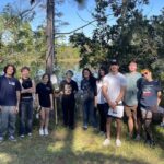 A group of twelve people stands outdoors on grass near trees and a lake, posing for a photo under a sunny, blue sky. Some hold papers or clipboards.