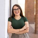 A woman with long brown hair and glasses stands smiling with arms crossed, wearing a green top and plaid skirt, leaning against a white wall with brick columns in the background.