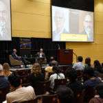 Two men sit on stage in armchairs, speaking to an audience during a lecture. Large screens display a headshot of one speaker and event information.
