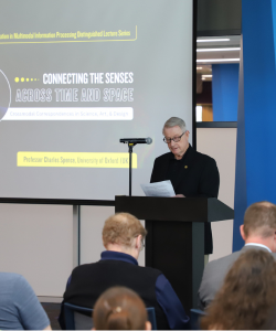 A man stands at a podium reading from papers during a lecture titled "Connecting the Senses Across Time and Space," with attendees seated and listening.