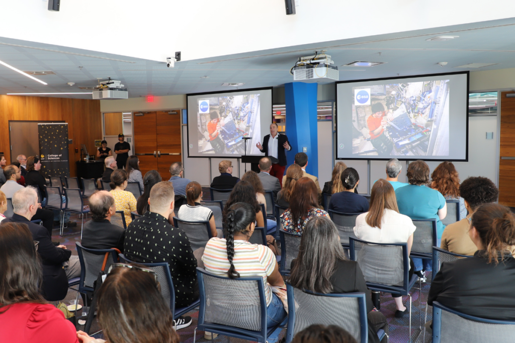 A speaker presents slides to an audience seated in a conference room, with images of astronauts on the screens at the front.