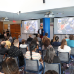 A speaker presents slides to an audience seated in a conference room, with images of astronauts on the screens at the front.