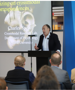 A man speaks at a podium in front of a projected presentation slide titled "Keynotizing on crossmodal correspondences" to an audience in a lecture hall.