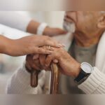 A caregiver places a reassuring hand on an elderly man’s hands as he holds a wooden cane, offering support and comfort.