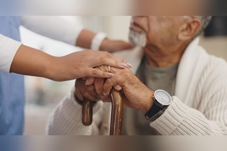 A caregiver places a reassuring hand on an elderly man’s hands as he holds a wooden cane, offering support and comfort.