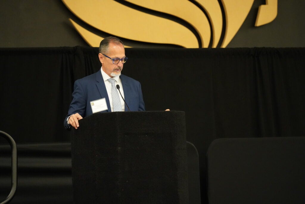 A man in a suit and glasses stands at a podium, speaking in front of a black curtain and a large gold and black abstract logo on the wall.