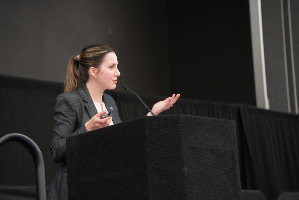 A woman in a suit stands at a podium, speaking into a microphone and gesturing with one hand during a presentation.