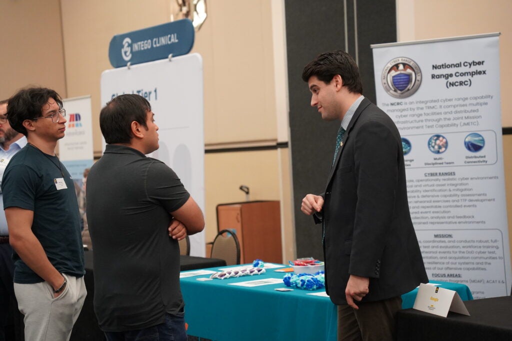 Three men stand and talk at a booth for the National Cyber Range Complex during a professional event or career fair. Pamphlets and informational materials are visible on the table.