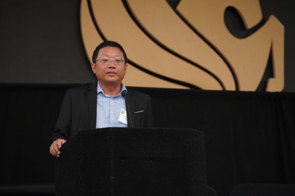 A man in a suit and glasses stands at a black podium, speaking in front of a large abstract logo on a black background.