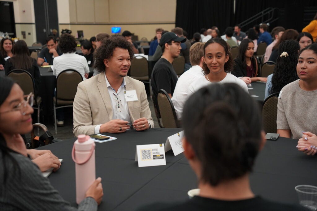 Several people sit around a round table, engaged in conversation at a professional event or conference in a large room with other groups in the background.