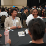 People sit around a round table having a discussion at a conference or networking event in a large room filled with attendees.
