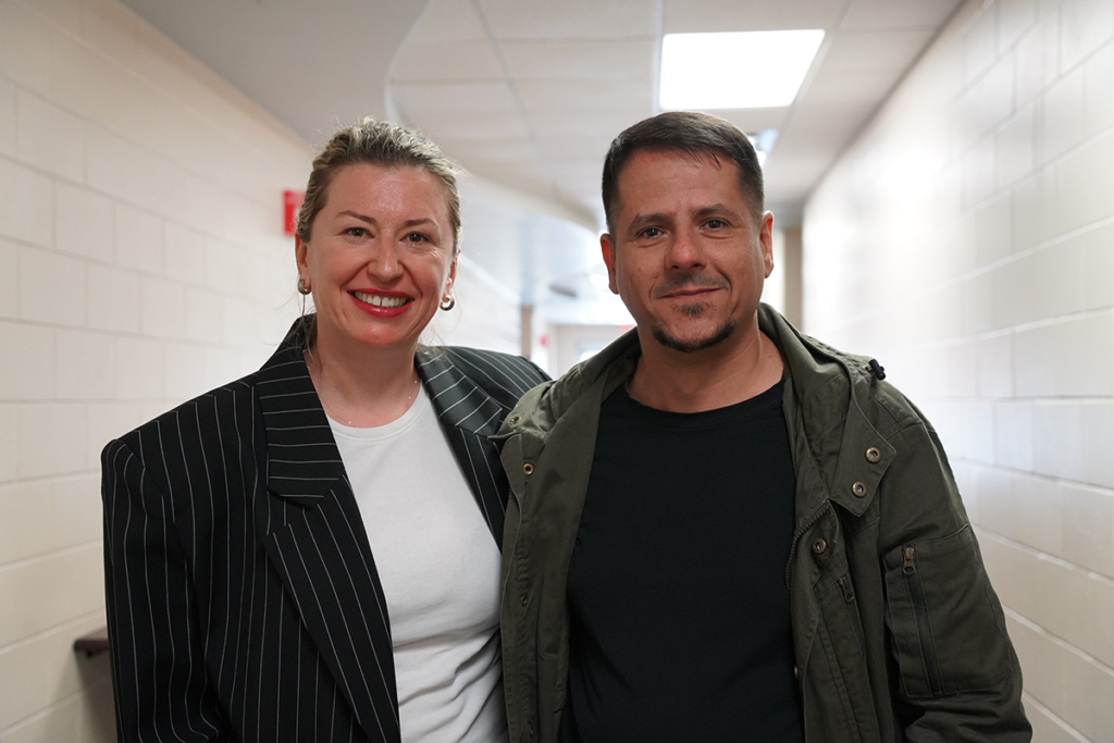 Two people stand side by side in a hallway with white brick walls and a ceiling light overhead, both looking at the camera and smiling.