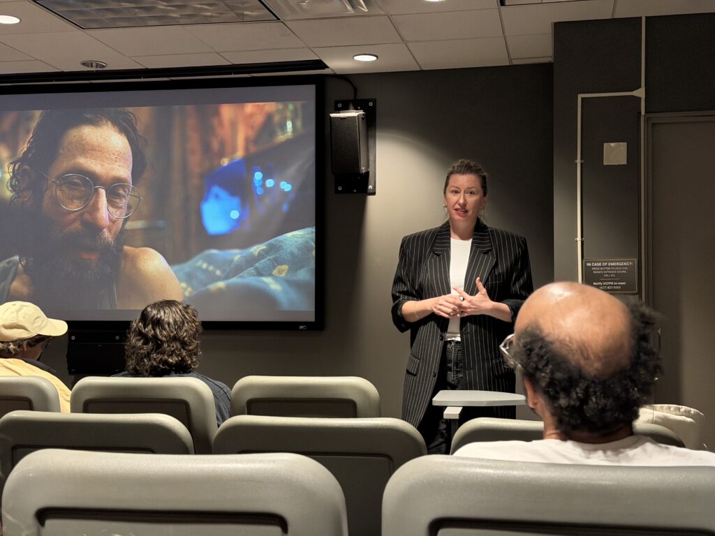 A woman stands and speaks in front of a small audience in a screening room, with a film scene featuring a man displayed on the screen behind her.