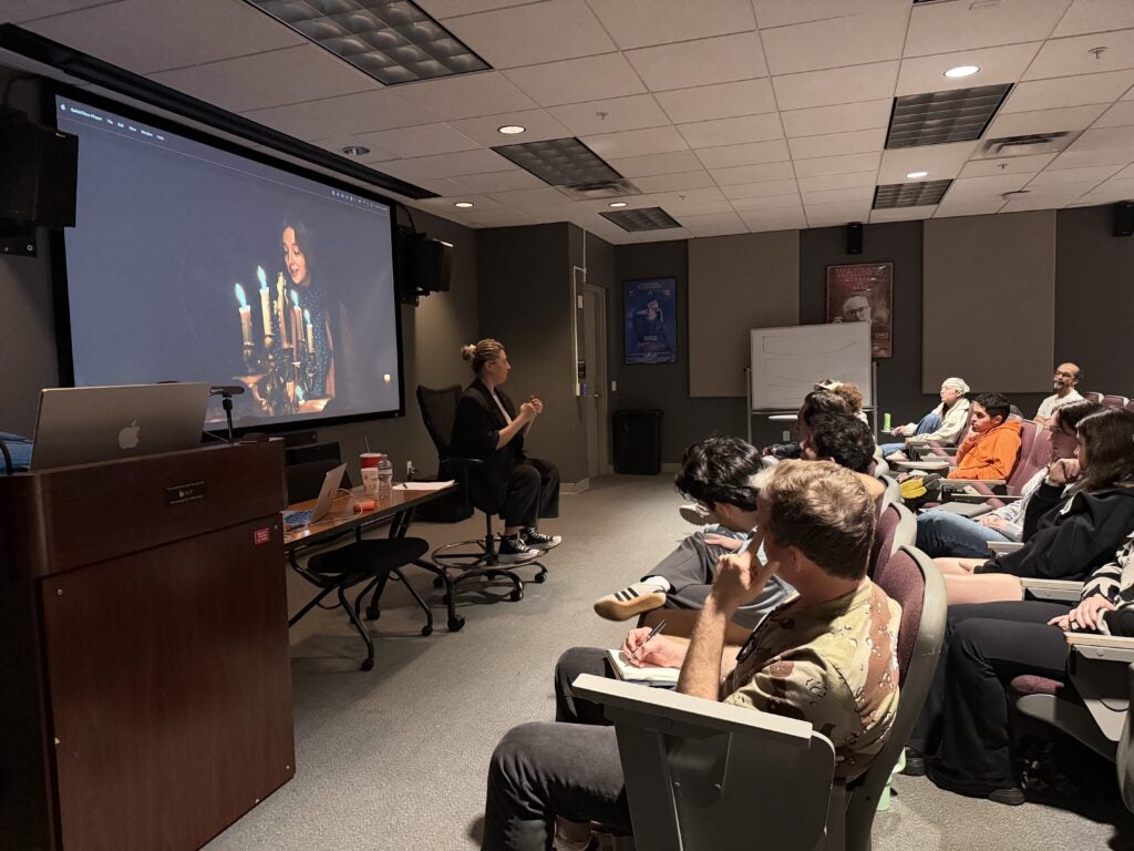 A woman gives a presentation to an audience in a classroom; a photo of a person with candles is projected on the screen behind her.