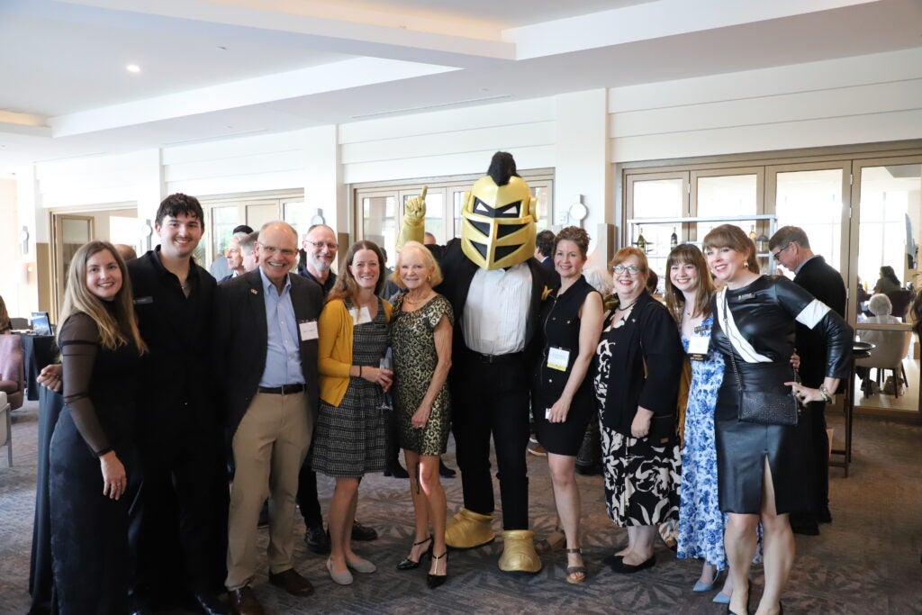A group of people stands indoors posing for a photo with a person in a knight mascot costume at an event.