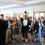 A group of people poses indoors with a person in a knight mascot costume, all smiling and dressed in business or semi-formal attire.