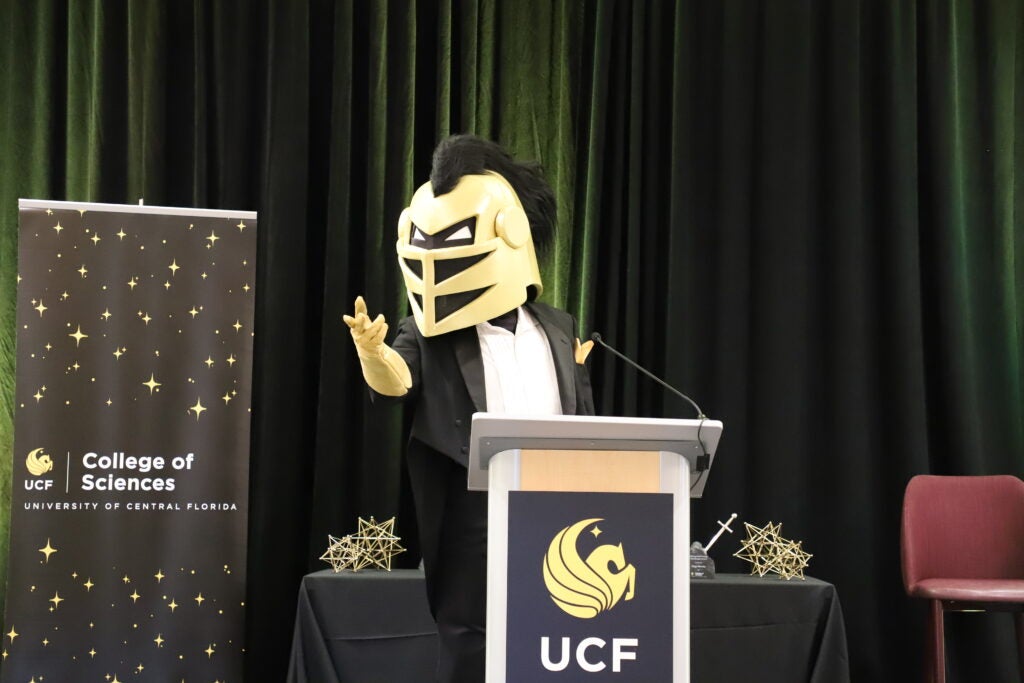 A person in a black and gold knight mascot costume stands at a podium with a UCF logo, next to a College of Sciences banner.