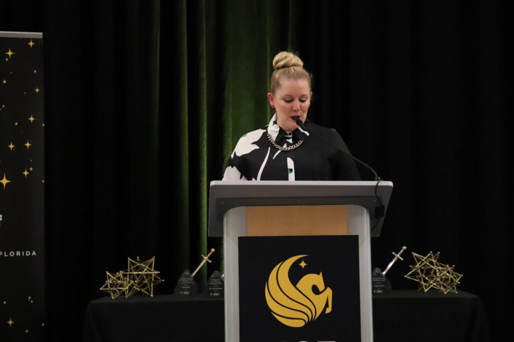 A woman stands at a podium with a microphone, speaking at an event. Geometric awards and a UCF logo are visible on the table behind her.