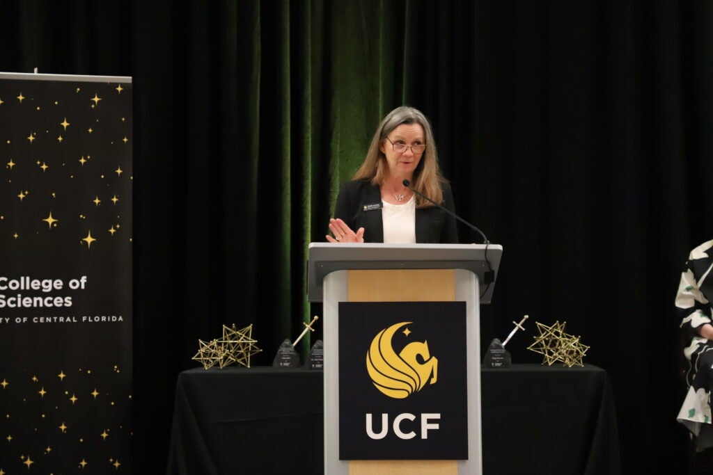 A woman stands at a podium with the UCF logo, speaking at an event. Behind her is a College of Sciences banner and a table with awards and geometric decorations.