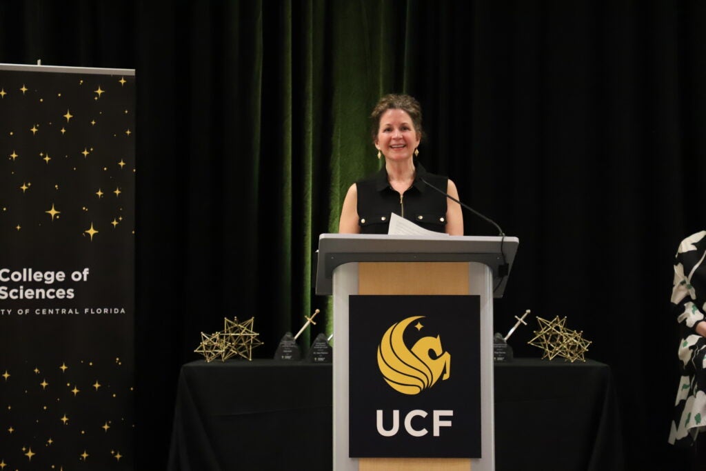 A woman stands at a podium with the UCF logo, speaking at an event. A College of Sciences banner and geometric awards are visible in the background.