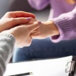 Two people sit close together, one gently holding the other's hand, with a clipboard and pen visible in the foreground.