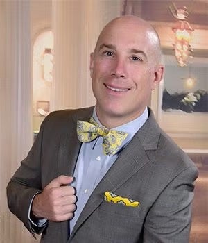 Bald man in a gray suit with a yellow patterned bow tie and matching pocket square, standing indoors and smiling at the camera.