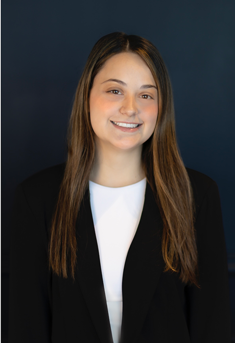 A young woman with straight brown hair, wearing a black blazer over a white top, stands and smiles in front of a plain dark blue background.