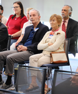 A group of adults seated in rows at an indoor event, attentively listening, with some holding their hands in their laps.