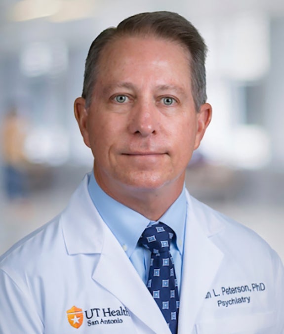 A man in a white lab coat with "UT Health San Antonio" and "Psychiatry" on it stands indoors, looking at the camera.